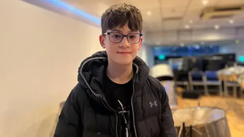 Pablo is wearing a black puffer coat and blue rimmed glasses behind steel pans.