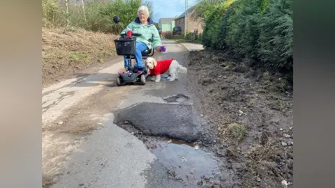 Pat rides her electric scooter that has a black basket on the front. She's wearing a green coat and blue trousers. Her small white dog, in a woolly red jumper, is on its lead next to her. They are on a country lane with houses in the background. There is a large, water-filled pothole in front of them.