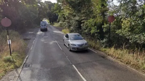 A Google Street View image of a country lane with a narrow footpath alongside one side of the road