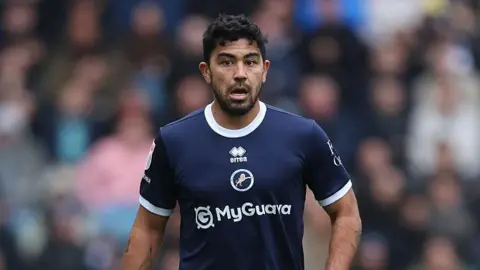 Massimo Luongo standing on the pitch wearing Millwall kit during a match in 2025