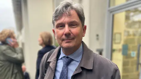 BBC Deputy Andy Sloan - A man with floppy grey and black hair looking at the camera. He is weariong a grey jacket over a blue shirt with a blue and white tie. 