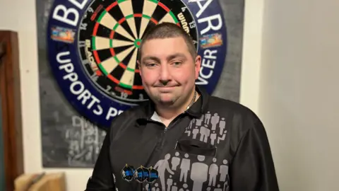 A man with black hair in a short buzz cut style. He is in a black darts shirt and holding three darts that have the Prostate Cancer UK logo on the flights. He is stood in front of a darts board.