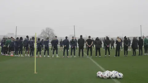 Oxford United A group of Oxford United's men's first team and academy held a minute's silence. They are stood around the centre circle heads bowed.
