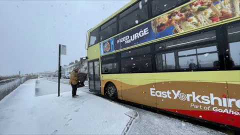 Woman at a bus stop with a scarf around her head. She is wearing a beige coat and black trousers and is closing her umbrella down as she stands in front of a bus that has just pulled up 