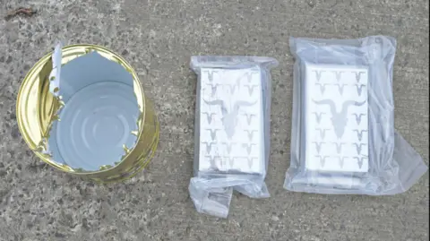 PSNI An opened, empty gold food can sits on the left while two rectangles of suspected drugs are wrapped in plastic on the right. All three items are placed on the concrete ground.