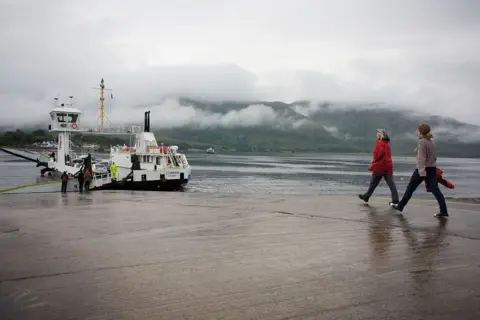 Two women, one wearing a red jacket and the other purple fleece, walk down a rain-soaked jetty towards the Corran Ferry service's MV Corran. The vessel is painted white and black. There are mist-shrouded hills on the opposite shore.