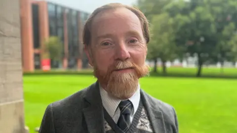 A man with a red-orange beard and flowing moustache wearing a patterned, knitted jumper and dark grey jacket looks at the camera.