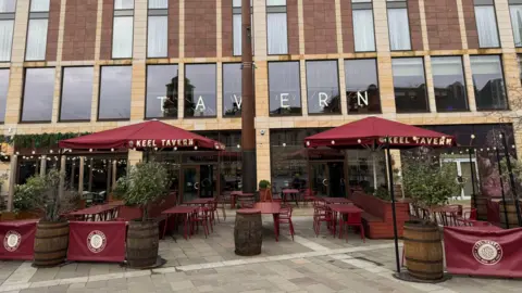 Outside of Keel Tavern bar. Large windows have lettering plastered on the insides which say "Tavern". Outside the building are red tables and parasols. The parasols say Keel Tavern