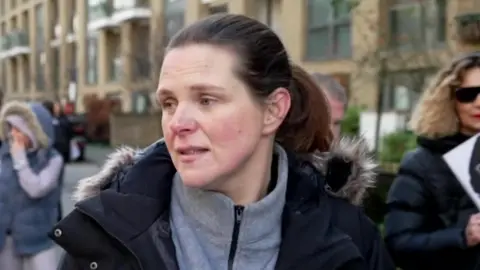 A woman in a grey jumper and black coat on a protest march. She has brown hair in a ponytail.