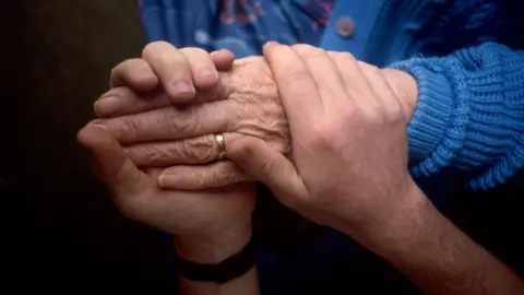 BBC Health carer holds the hand of an elderly patient.