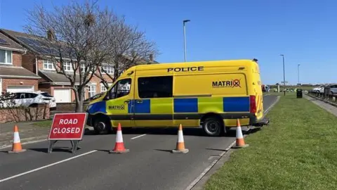 A yellow Cleveland Police Matrix van is parked across a road behind a road closed sign and traffic cones.
