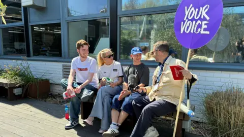 Zac Sherratt/BBC Left to right, Samuel, 21, Caitlin, 21, and Taylor, 20, who are all graphics students are interviewed by BBC South East Today reporter, Peter Whittlesea. Peter holds a sign saying Your Voice. They are all sat on a bench.