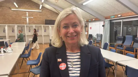 Helen Miles, a woman with shoulder length blonde hair and white earrings. She is wearing a white and navy stripy top underneath a navy blazer. On her lapel is a badge with a Jersey flag, and a sticker saying 'I'm a voter'. A number of tables are visible behind her in a church hall.