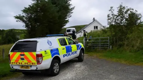 Two police vehicles and two officers outside a white single-storey house in a rural area