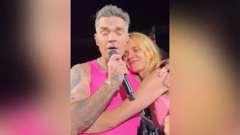 Liga Bri A woman with blonde hair cuddles up to Robbie Williams and has her eyes closed in glee with a smile on her face. Williams wears a pink vest and has his eyes closed as he sings into a microphone