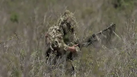 A British sniper wearing camouflage and a helmet covered in foliage takes part in training while in Kenya