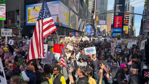 Grace Eliza Goodwin/BBC image of thousands of protestors gathered in street, holding signs, with a big American flag waving