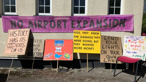 Homemade protest signs are leaning against a wall. One of them says Keep The Airport Off Felton Common. Another says History Has Its Eyes On Us.