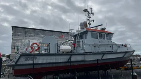 BBC A grey ship, The Leopardess, behind a fence. It has the States of Guernsey logo on the side. 