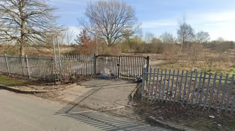 Google Derelict land, including a concrete roadway and grassland, is guarded by metal fencing and black double gates. A number of tall bare trees stand at intervals against a wintry blue sky. 