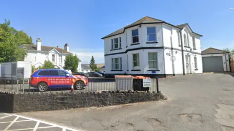 Google A large white two-storey building, a driveway and a red and blue car.