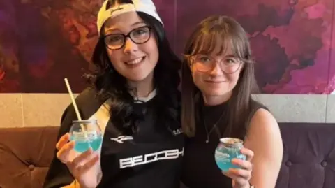 Caitlin and Katy sit on a purple sofa smiling while holding light blue BuzzBall canned cocktail drinks.