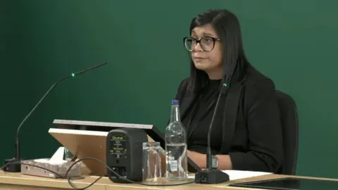 A woman sat at a desk in front of a screen and two microphones. She is wearing black clothing, has dark hair and black-rimmed glasses.