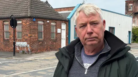 A man in his early 60s, with short, grey hair, looks at the camera. He is wearing an emerald green jacket over a grey fleece and blue t-shirt. Behind him is Reform Street. 