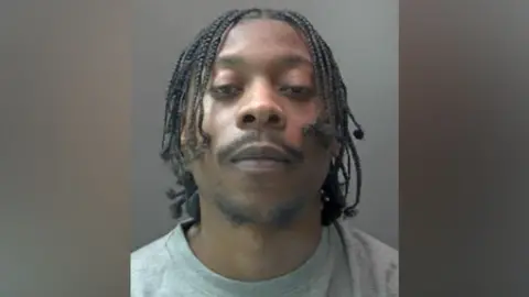 Hertfordshire Police A police custody image of a defendant. He has slightly raised his head up and is looking directly at the camera. He is expressionless and has his eyes half closed. He has a dark, wispy, goatee beard and a moustache. His hair hangs in small plaits.