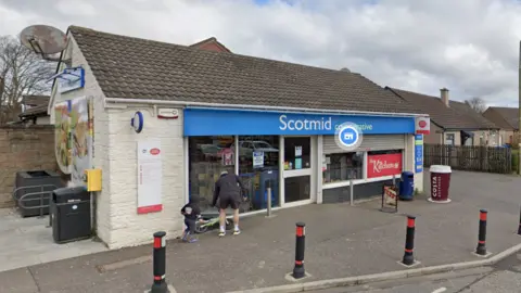 The front of the Scotmid shop has a a sign in blue. There are five bollards along the edge of the pavement in front of the shop.