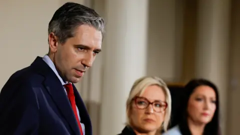 Reuters Simon Harris, wearing a suit, blue shirt and red tie, to the left of the photo. Michelle O'Neill and Emma Little-Pengelly are in the blurred background of the photo. 