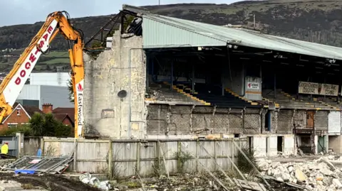 Rubble and bricks are lying on the grass as a yellow digger is knocking down a grandstand.