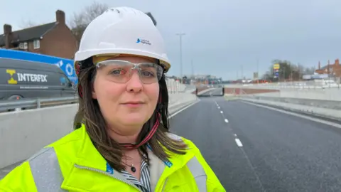 Shot of a woman wearing safety googles, hard hat and a yellow high-vis jacket. She is standing close to where the road leads down to an underpass.