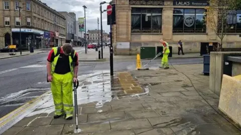 Two men in hi-vis clothing using power washers to clean streets of gum.