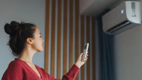 A woman with brown hair, wearing a red cardigan, is turning on a wall-mounted air-conditioning unit with a remote.