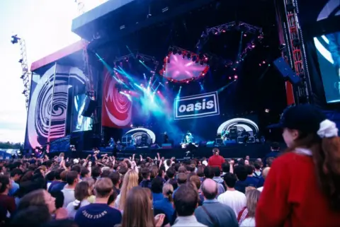Getty Images A shot of Oasis on stage at an outdoor concert, shot from the middle of the audience