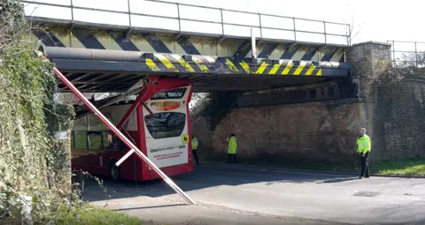 A red and white Pulhams bus is lodged beneath a railway bridge on a country lane. The roof of it looks to have been damagd and partially ripped off. Police officers are stood beneath the bridge with the bus, wearing hi-vis jackets. What appears to be part of the roof of the bus - resembling a long pole - is propped up against the bridge.