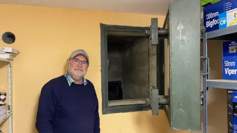 Edd Smith/BBC Paul Hendry-Smith wearing a cap, glasses and navy jumper over a shirt, smiling while standing in front of a yellow coloured wall on which there is a large concrete and metal square door which is open and leads to an escape tunnel. 