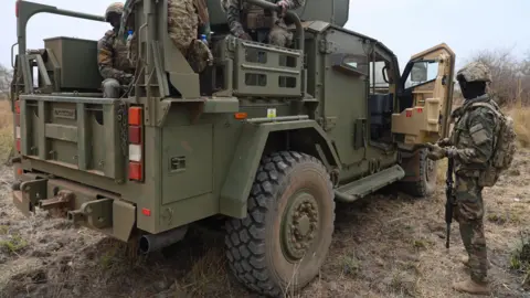 Ghanaian soldiers in camouflage uniform on a green military truck with the front door open and one soldier standing beside it 