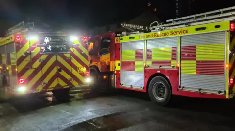 Aaron Chesham The backs of two red and yellow fire engines at the scene. It is night time and they have their lights on. The side of the vehicle reads 'fire and rescue service'.