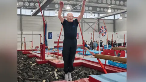Sue Burt Quinney standing on the edge of a trampoline, balancing on his tip toes with his arms stretched up, looking as thought he is about to start a routine. He is wearing a navy top and dark sports trousers. Behind him is a gymnastics sponge pit and in the background are various bits of gymnastics equipment including beams. There are also union jack flags.