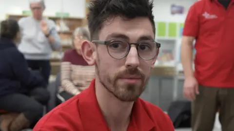 Jamie Niblock/BBC Josh Lawrence, a man who is looking directly at the camera. He is wearing a red polo shirt and round glasses.