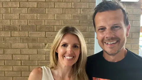 Ollie Conopo/BBC Jaime Thurston, who has long blonde hair, is smiling and standing to the left of Greig Trout, who has short brown hair and is also smiling. They are standing in front of a brick wall.