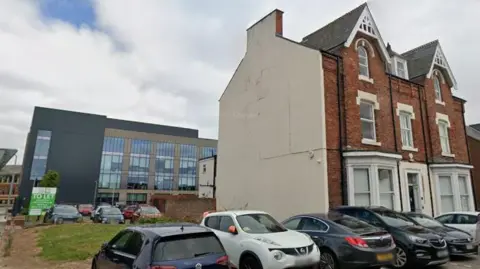 Google Cars parked outside a terraced house and a patch of land to the right. In the background there is a large, modern, glass building. 