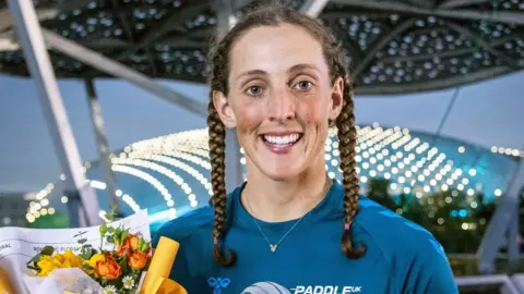A close-up image of a woman with brown hair in two plaits, wearing a blue t shirt and smiling at the camera