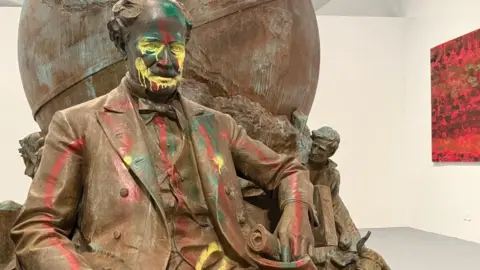 BBC / Regan Morris A bronze statue of an old-fashioned dressed man, sitting in front of a bronze globe, is covered in graffiti