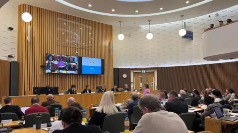 BBC Round conference room with wooden panels on walls, people seated behind long tables with microphones, facing a wooden panel of people at front with screen behind them screening the meeting