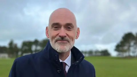 Professor Peter Bradley is wearing a navy overcoat with the collar turned up. He has a silver beard and is looking directly at the camera.