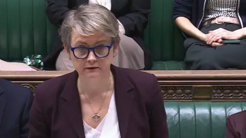 Yvette Cooper delivering a speech in the House of Commons wearing large glasses with blue frames, a jacket and white blouse with a silver pendant chain around her neck