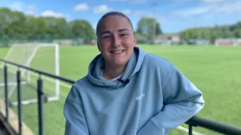 Chloe is standing on the side lines of a football pitch wearing a blue hoodie, her hair is tied back and she is smiling at the camera. There is a football goal in the background.